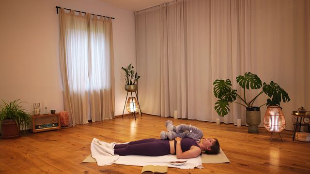 Isabell liegt in einem warm beleuchteten Raum auf der Yogamatte, auf ihrem Bauch liegt Eli, die Elefantin, als Kuscheltier