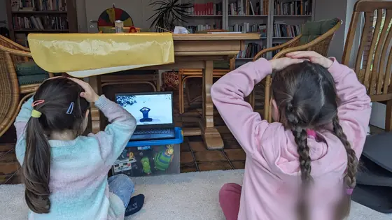 Zwei Mädchen sitzen vor dem Laptop und machen mit den Händen am Kopf eine Übung nach
